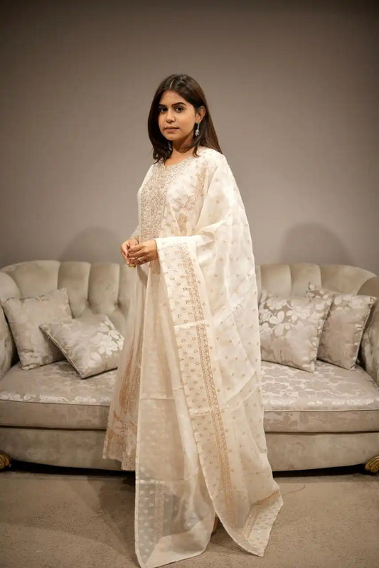 Woman in a white traditional outfit standing in a room with a beige sofa and decorative pillows.