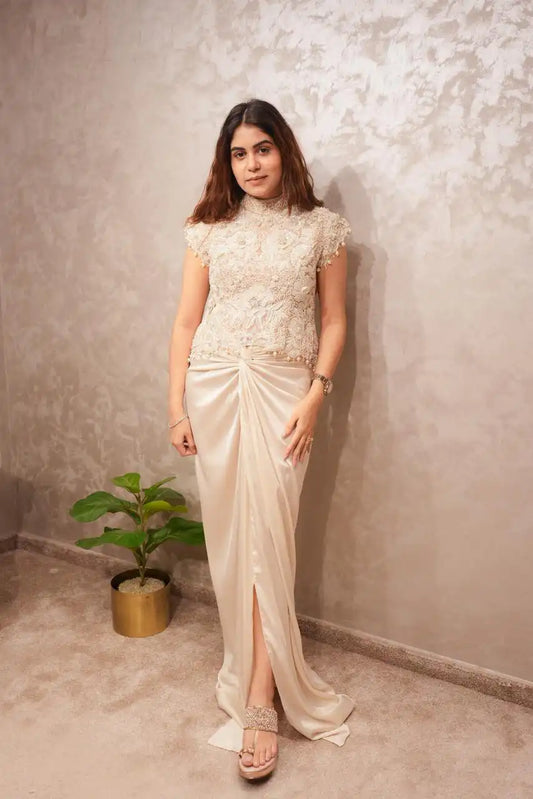 Woman in a white floral dress standing against a textured wall with a plant beside her.