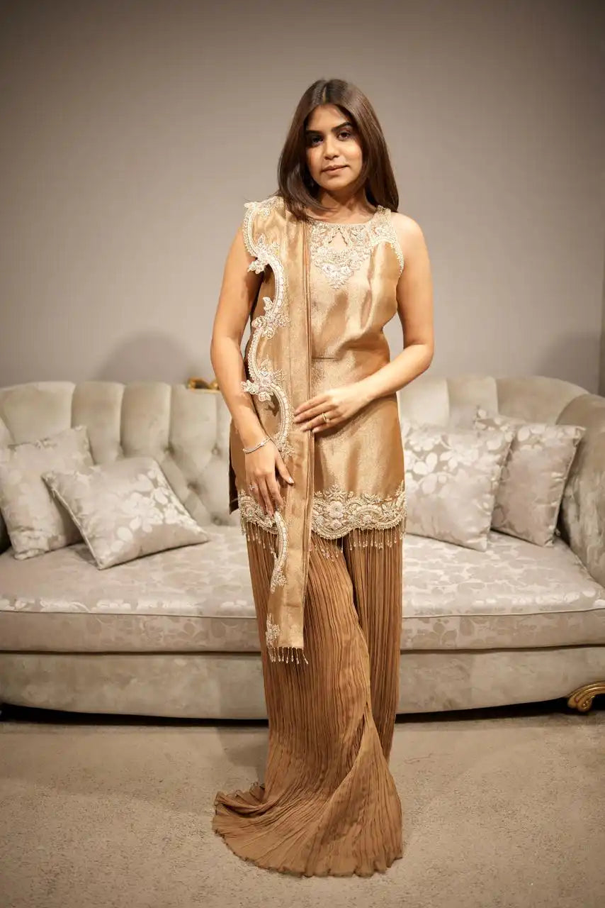Woman in a beige saree with a matching blouse standing in front of a gray sofa.