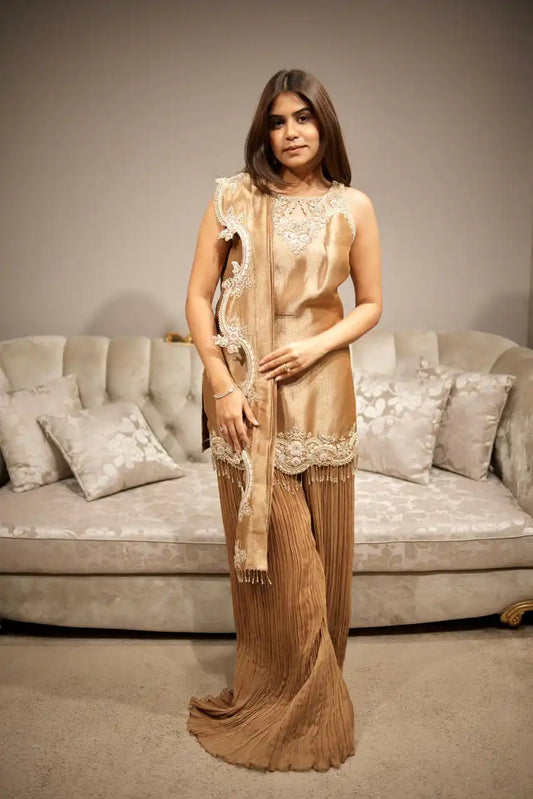 Woman in a beige saree with a matching blouse standing in front of a gray sofa.