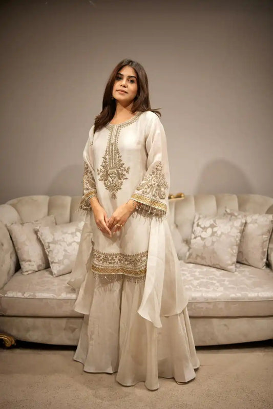 Woman in an embroidered traditional outfit standing in a living room with a sofa and decorative pillows.